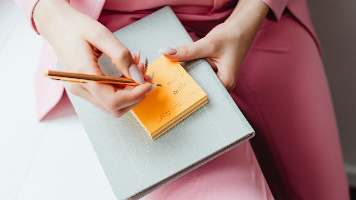 Close-up of a woman writing on a sticky note with a pencil, wearing a pink suit.